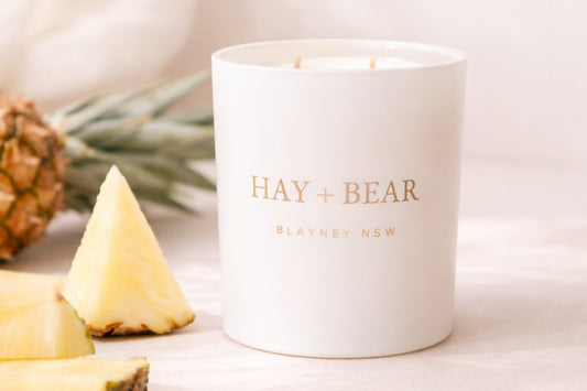 White candle with 'Hay + Bear' branding next to pineapple slices on a light background